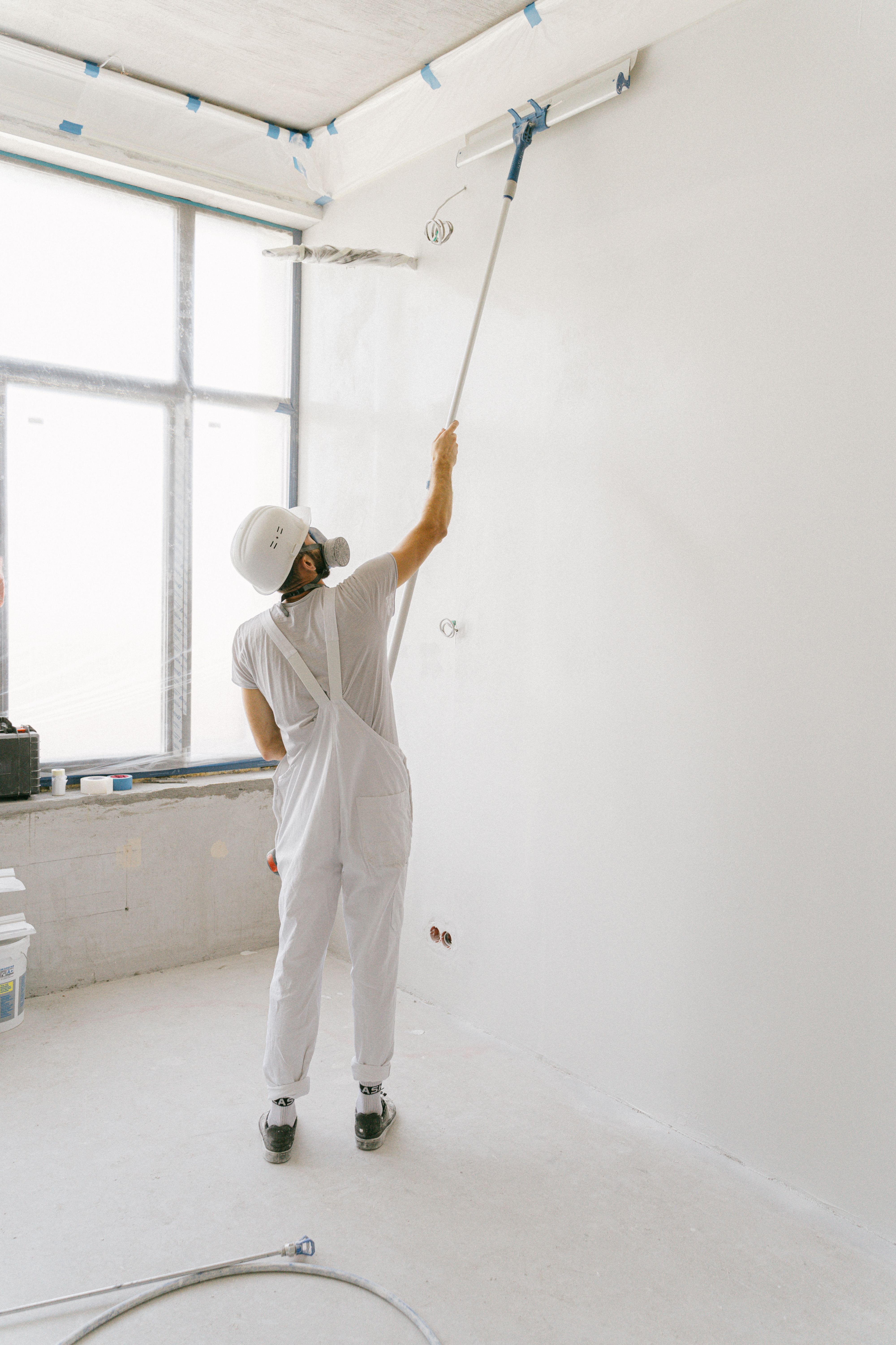 Artisan peintre en équipement blanc appliquant une peinture soignée au rouleau perche près d'une grande fenêtre